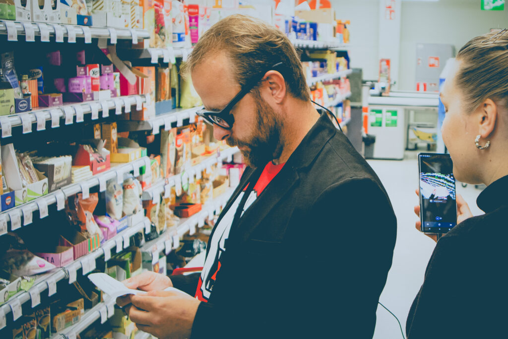 Käytettävyyslaboratorion testaustilanne, jossa henkilö kaupassa hylkyjen edessä, silmillään käytettävyystestaukseen tarkoitetut silmälasit.